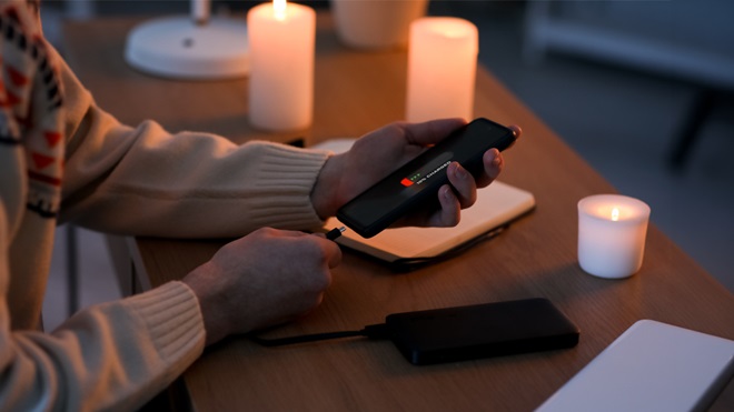 Persona che ricarica uno smartphone con un power bank durante un blackout, illuminata da candele su un tavolo di casa.