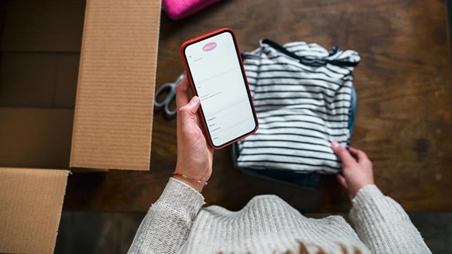 mano di donna tiene telefono in mano con app aperta con dietro scatolone da riempire con vestiti